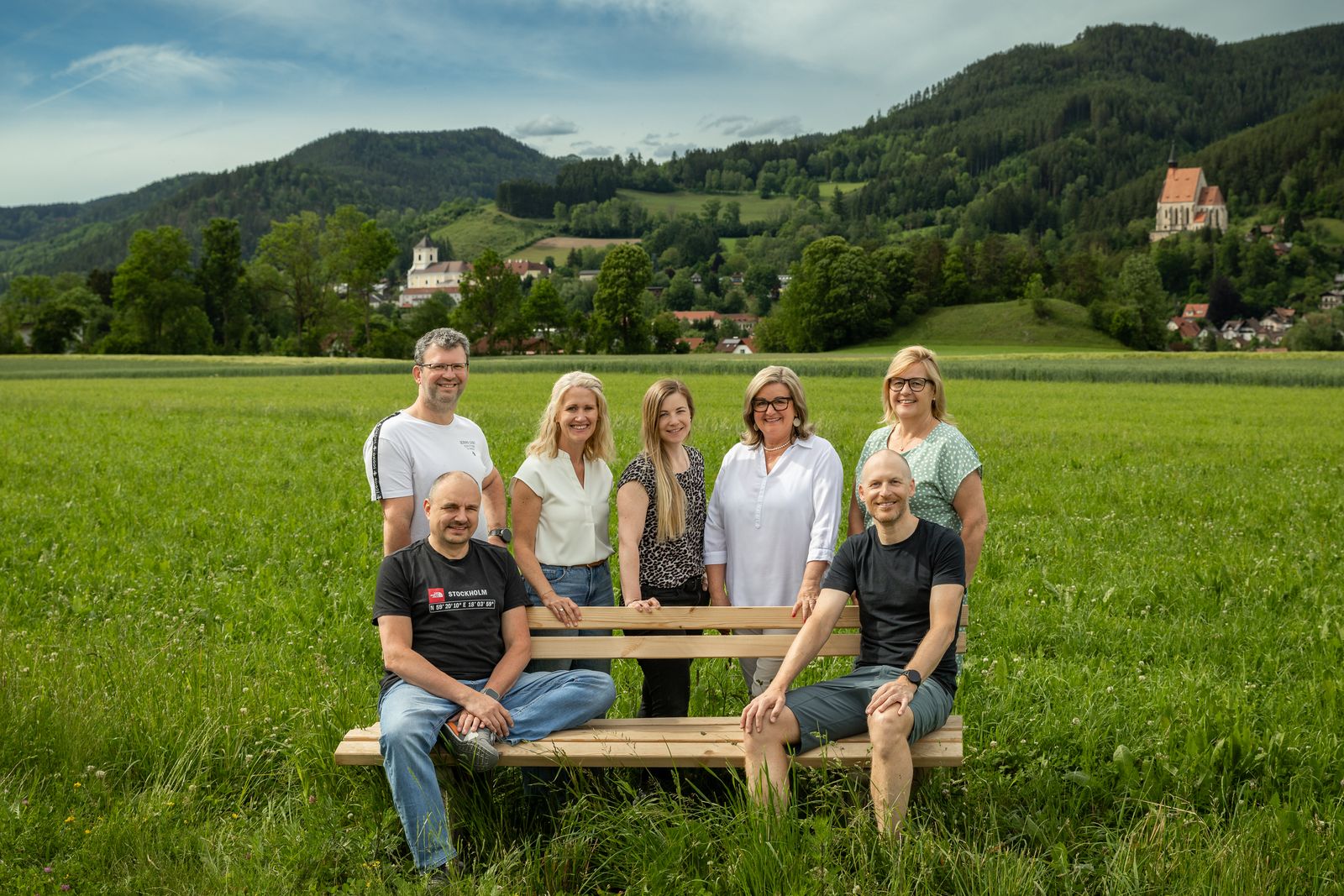 Gruppe auf einer Holzbank in der Wiese, im Hintergrund Kirchberg am Wechsel mit Pfarrkirche und St. Wolfgangskirche